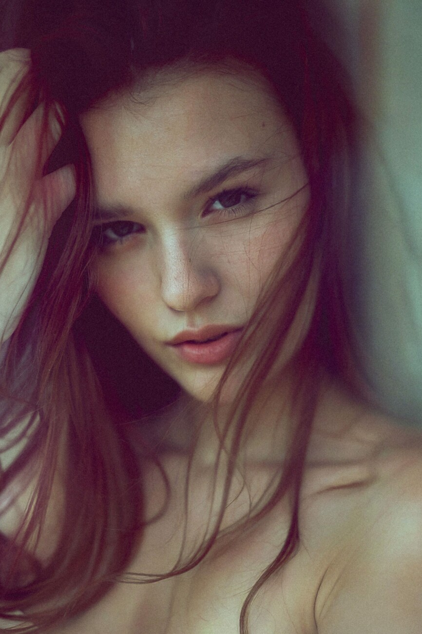 Close-up of a young woman with long hair, touching her face and looking softly at the camera
