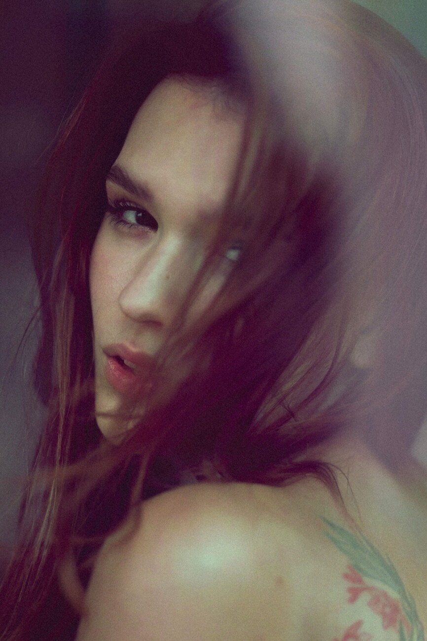 Close-up portrait of a person with long hair partially covering the face, soft lighting and a pastel floral tattoo on the shoulder visible.