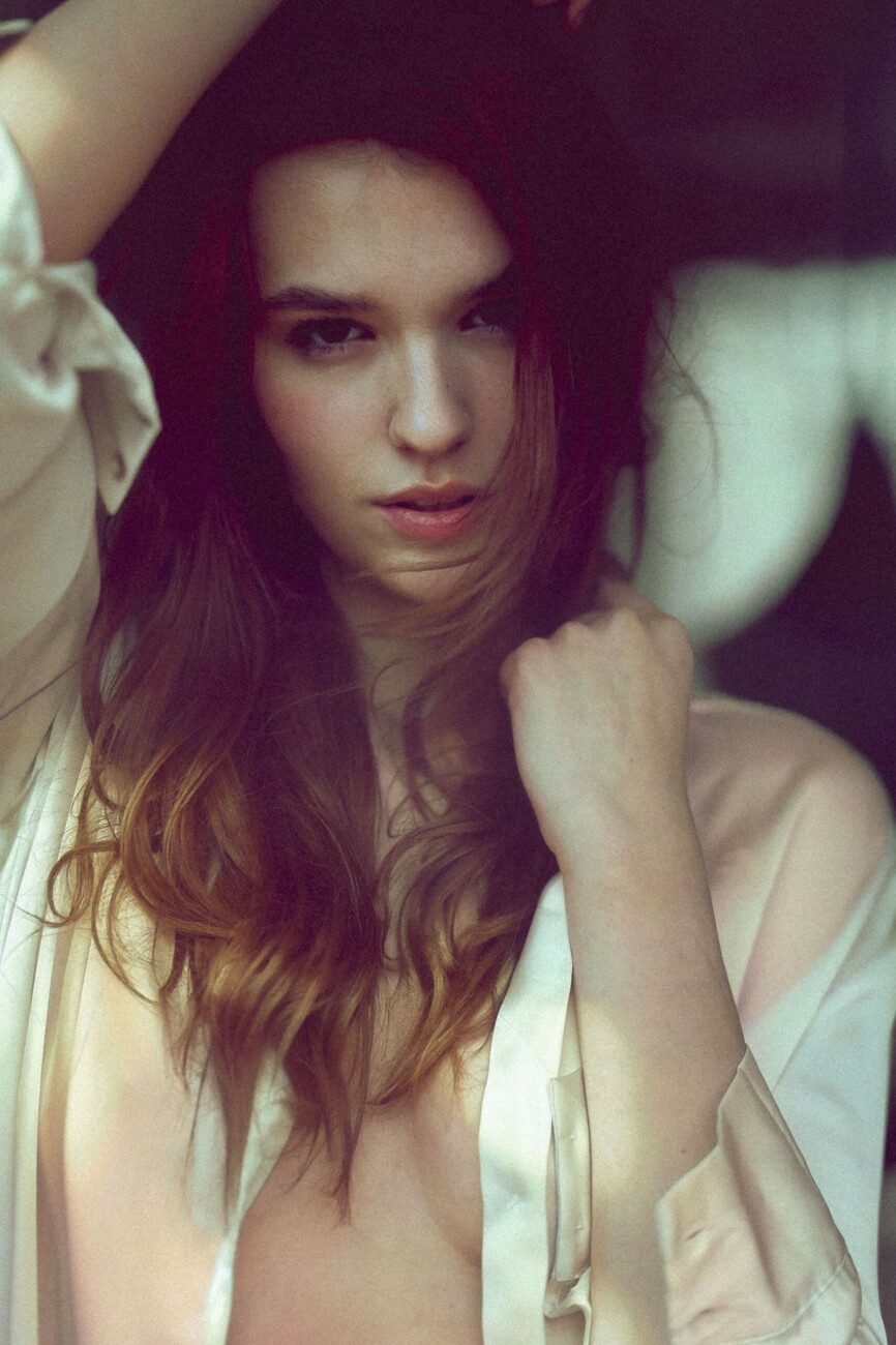 Close-up of a woman with long hair in a pale satin shirt, looking at the camera.