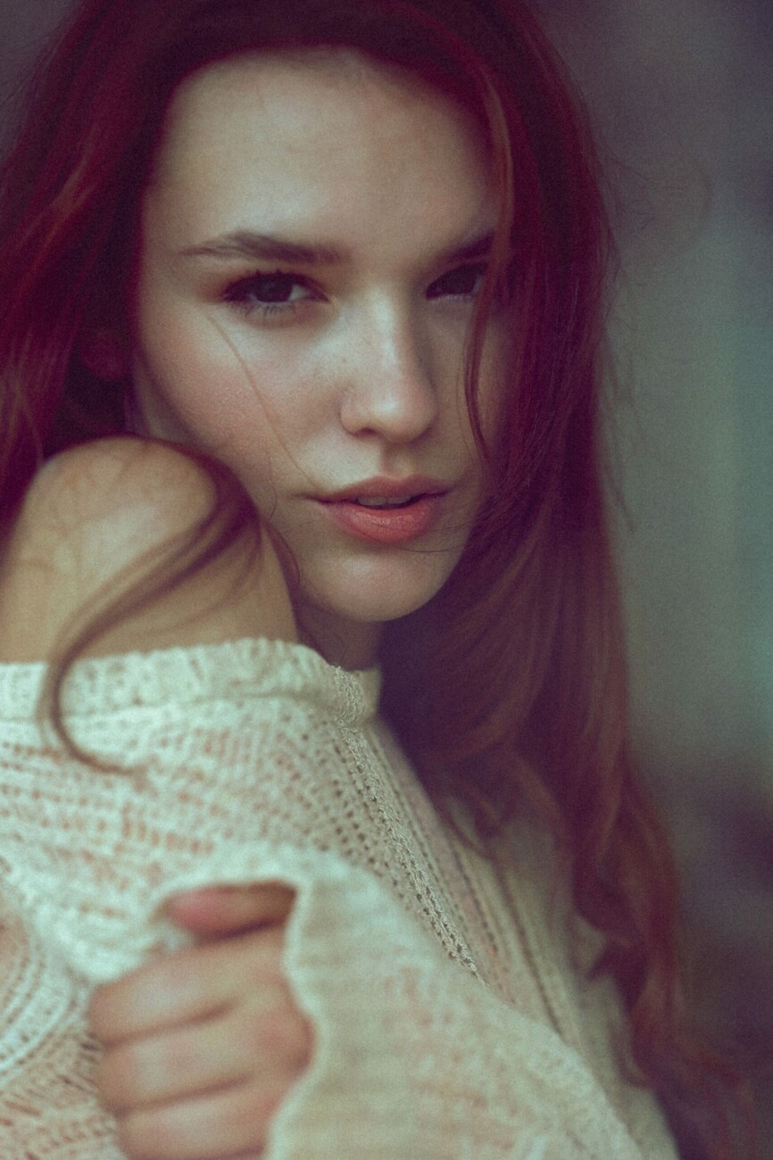 Close-up portrait of a woman with red hair resting her chin on her hand, wearing a cream knit sweater.