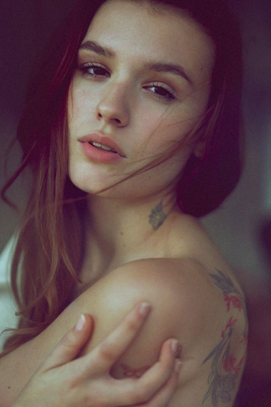 Close-up portrait of a woman with red hair, soft makeup, bare shoulders, and a floral tattoo on her shoulder.